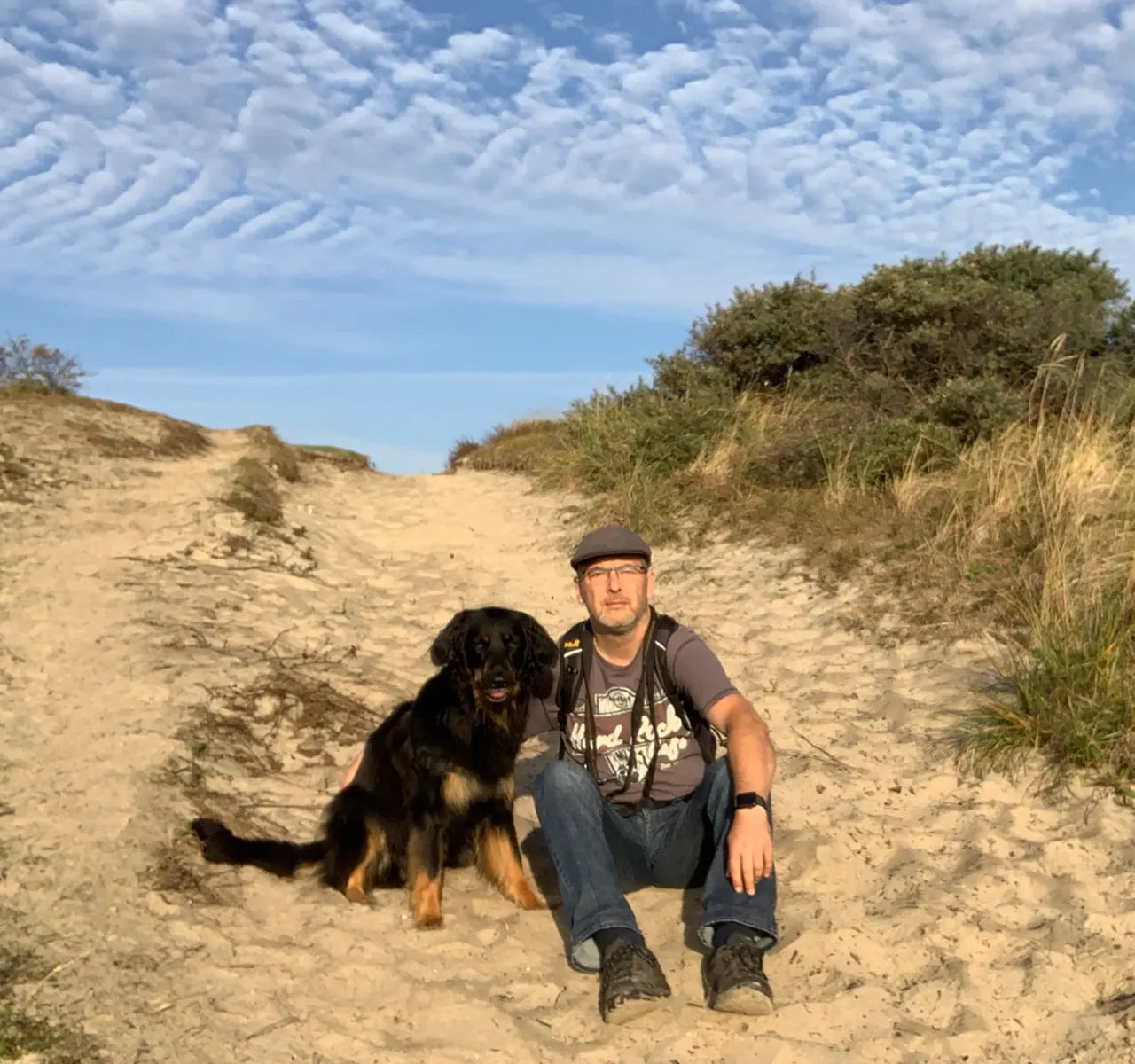 Alex von hovawart.rocks sitzt gemeinsam mit seiner schwarzmarkenen Hovawarthündin entspannt in einer Dünenlandschaft unter blauem Himmel.