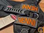 Nahaufnahme eines rechteckigen, stabil bestickten Patches in Schwarz. Auf dem Patch steht in kontrastreicher, orangner Schrift der Name "Henni". Der Patch hat eine Klettverschluss-Rückseite und ist für Hundegeschirre geeignet.
