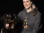 Lächelnde Frau in einem anthrazitfarbenen "Powerrun" Hovawart Hoodie mit großem, farbigem Hovawart Schwarzmarken Motiv auf der Brust, posiert neben einem realen Hovawart mit heraushängender Zunge vor schwarzem Hintergrund.