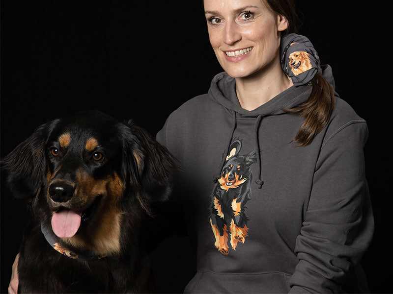 Lächelnde Frau in einem anthrazitfarbenen "Powerrun" Hovawart Hoodie mit großem, farbigem Hovawart Schwarzmarken Motiv auf der Brust, posiert neben einem realen Hovawart mit heraushängender Zunge vor schwarzem Hintergrund.