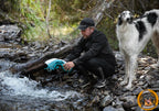 Ein Mann kniet an einem steinigen Bachlauf und wässert eine blau-weiße Kühlweste für seinen daneben stehenden schwarz-weißen Borzoi.