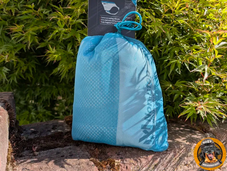 Eine blaue, durchscheinende Netzstofftasche mit Kordelzug, in der eine Hunde-Kühlweste verstaut ist, steht auf einem rauen Untergrund vor grünem Laub. Oben ist ein schwarzes Produktschild sichtbar. Unten rechts ist das runde Logo "Born to Rock - www.hovawart.rocks" eingeblendet.