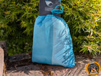 Eine blaue, durchscheinende Netzstofftasche mit Kordelzug, in der eine Hunde-Kühlweste verstaut ist, steht auf einem rauen Untergrund vor grünem Laub. Oben ist ein schwarzes Produktschild sichtbar. Unten rechts ist das runde Logo "Born to Rock - www.hovawart.rocks" eingeblendet.