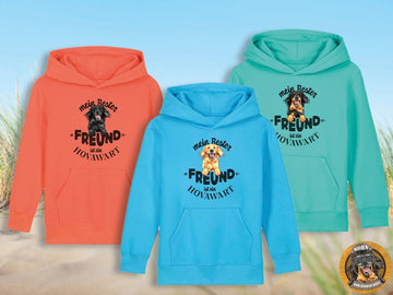 Drei Kinder Hoodies liegen nebeneinander auf einem sandigen Untergrund vor Strandgräsern und blauem Himmel. Der linke Hoodie ist in leuchtendem Orange (Fiesta) mit einem schwarzen Hovawart-Aufdruck. Der mittlere Hoodie ist in Aqua Blue mit einem blonden Hovawart-Aufdruck. Der rechte Hoodie ist in Pool Blue mit einem schwarz-markenen Hovawart-Aufdruck. Alle Hoodies zeigen den Text "mein Bester FREUND ist ein Hovawart" und haben eine Kängurutasche. Unten rechts ist ein rundes Logo mit einem Hovawart zu sehen.