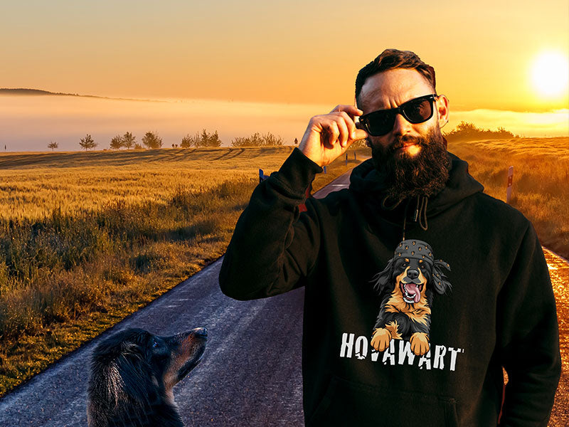 Mann mit Vollbart und Sonnenbrille trägt den schwarzen "Hovawart Rockstar" Hoodie mit farbigem Motiv eines Hovawarts auf der Brust. Er steht auf einer leeren Straße bei Sonnenuntergang neben einem realen Hovawart.