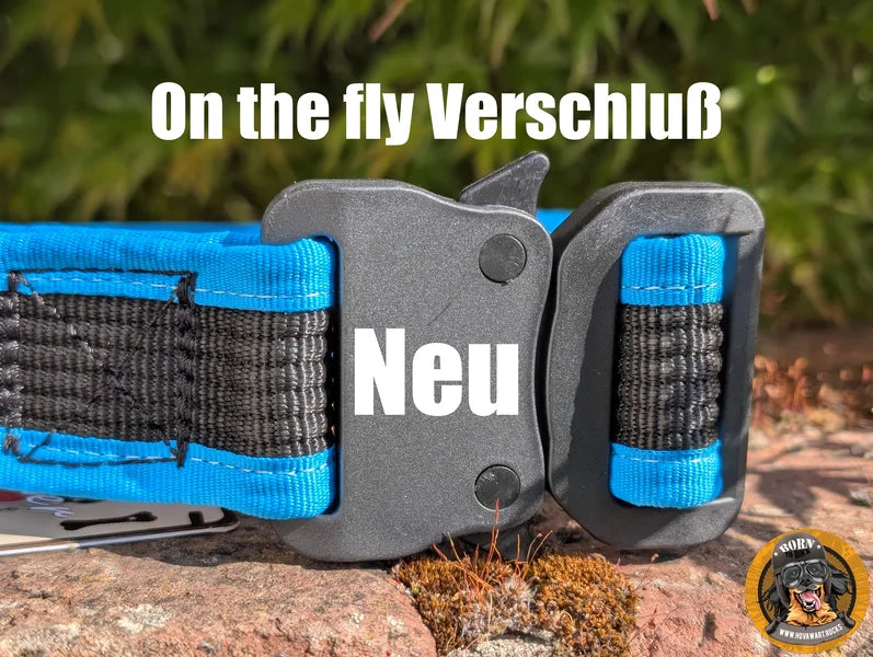 Nahaufnahme eines stabilen, dunkelgrauen Verschlusses an einem blauen und schwarzen Hundehalsband. Der Verschluss ist ein Schnellverschluss mit einem seitlichen Hebel. Auf dem oberen Teil des Bildes steht in weißer Schrift "On the fly Verschluß", und auf dem Verschluss selbst steht in weißer Schrift "Neu". Das Halsband liegt auf einem moosbewachsenen Untergrund. Unten rechts ist das runde Logo "Born to Rock - www.hovawart.rocks" eingeblendet.