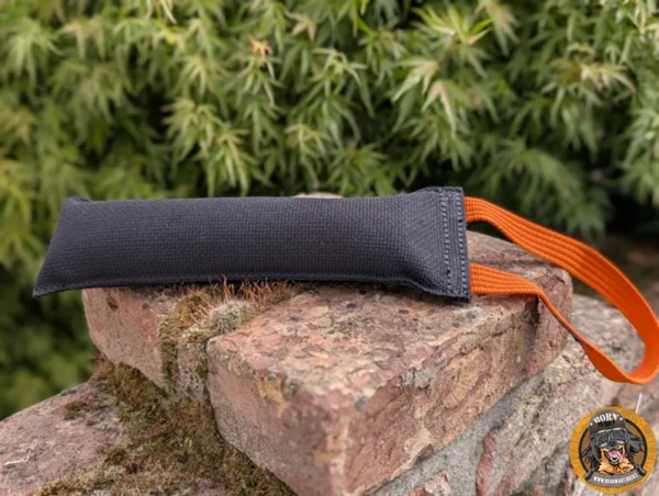 Grossenbacher Agility Boudin Zerrspielzeug mit oranger Schlaufe aus schwarzem Stoff, 27x8cm, auf Ziegelsteinmauer liegend