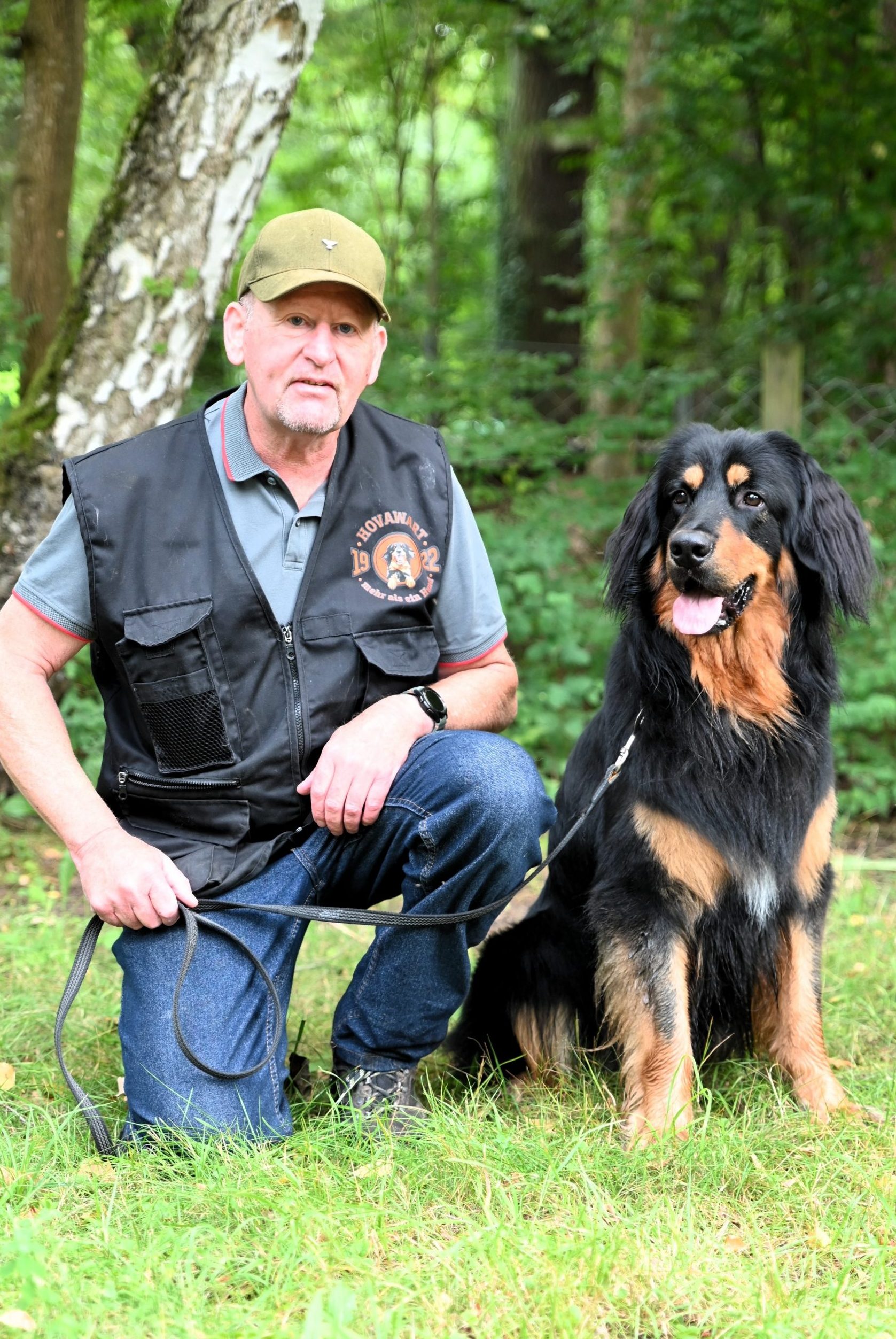 Hundesportler in schwarzer hovawart.rocks-Trainingsweste kniet neben einem wunderschönen schwarzmarkenen Hovawart auf einer grünen Wiese im Wald.